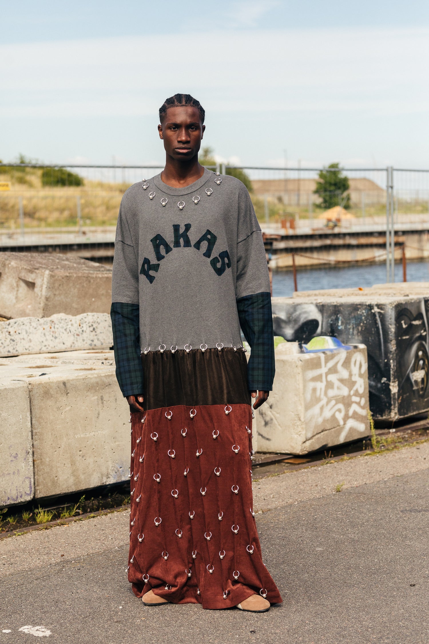 Mallilla on yllään mekko, jossa lukee isolla RAKAS. Mekossa on harmaa yläosa ja ruskeansävyinen alaosa. Asuun on kiinnitetty yli sata RAKAS Rolf Ekroth x Kalevala -korua.