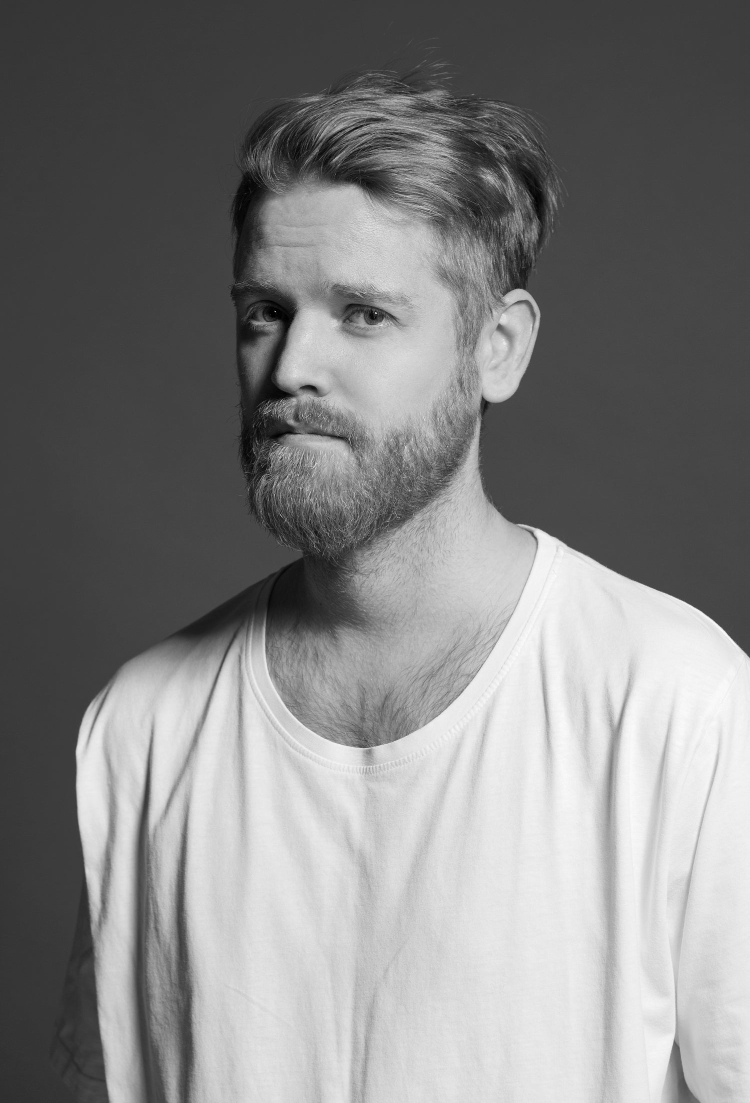 Muotisuunnittelija Rolf Ekroth katsoo vakavana kameraan mustavalkokuvassa. Hänellä on parta ja yllään valkoinen t-paita.